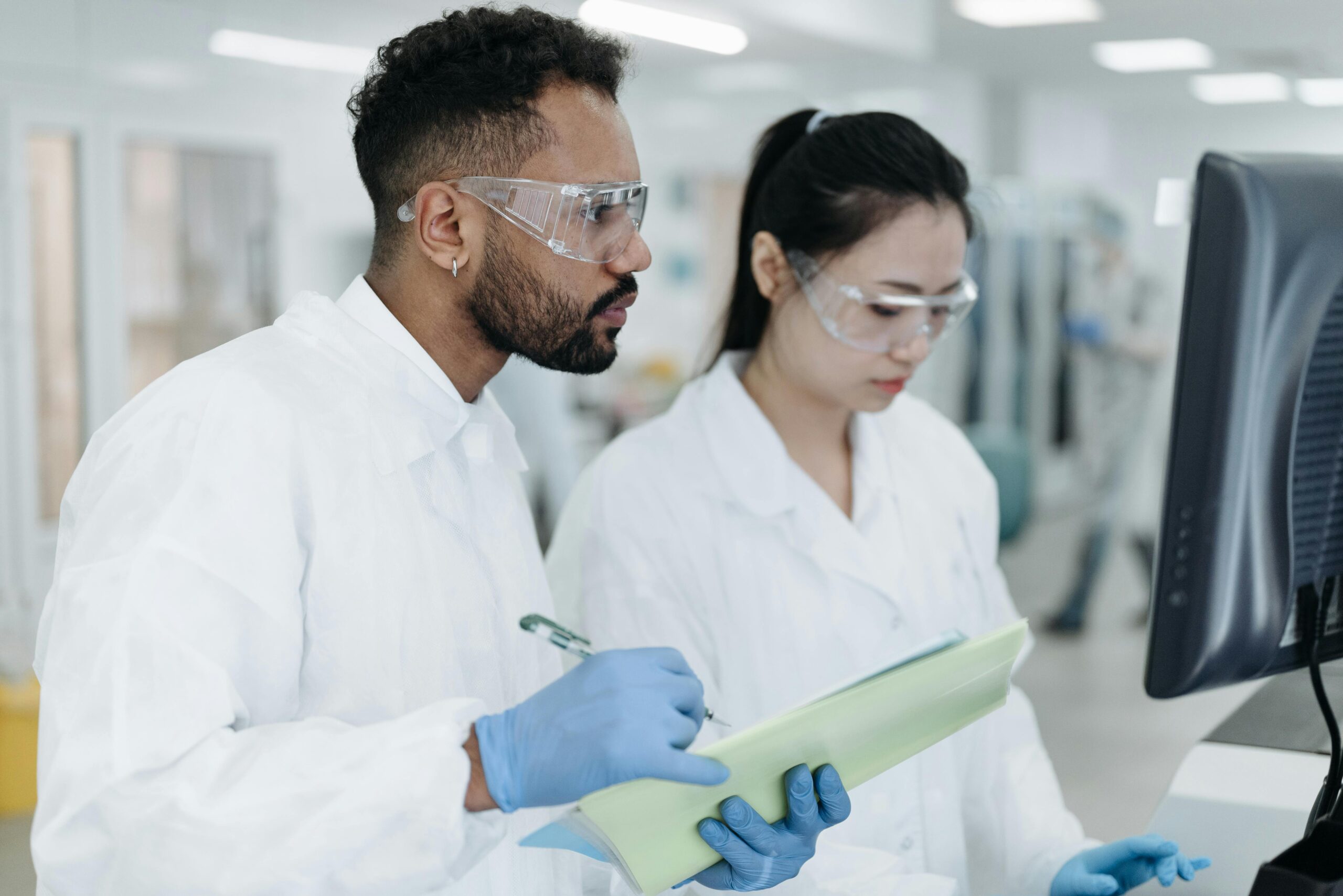 Two scientists in lab coats and safety goggles analyzing data at a computer in a modern biotech laboratory